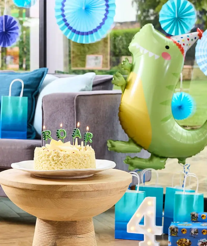 Dinosaur-themed birthday party display with a birthday cake, dinosaur balloon, blue decorations, gift bags, and a lit number 4 decoration.